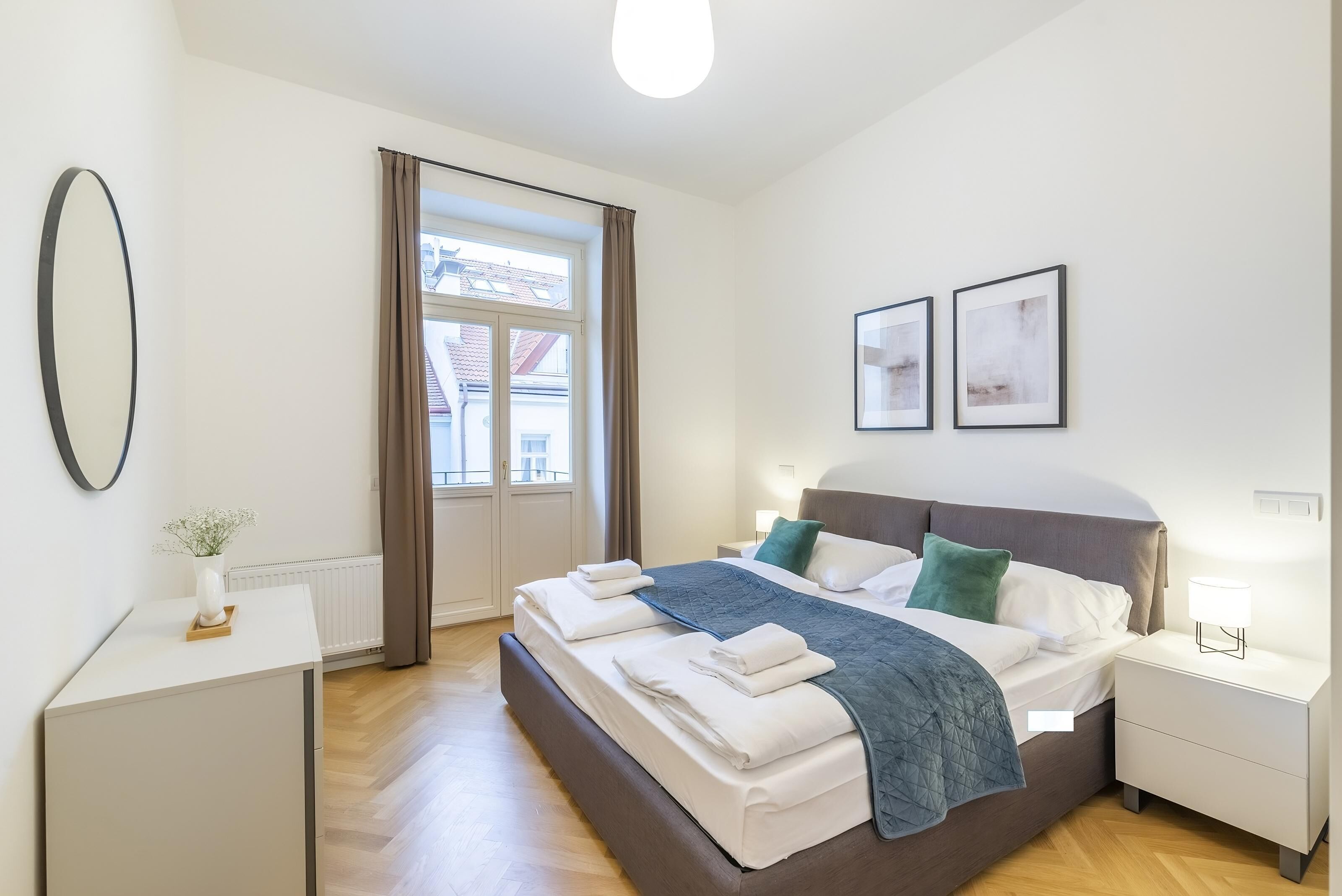 A modern bedroom with a large bed, bedside tables, a window with curtains, and framed pictures on the wall.