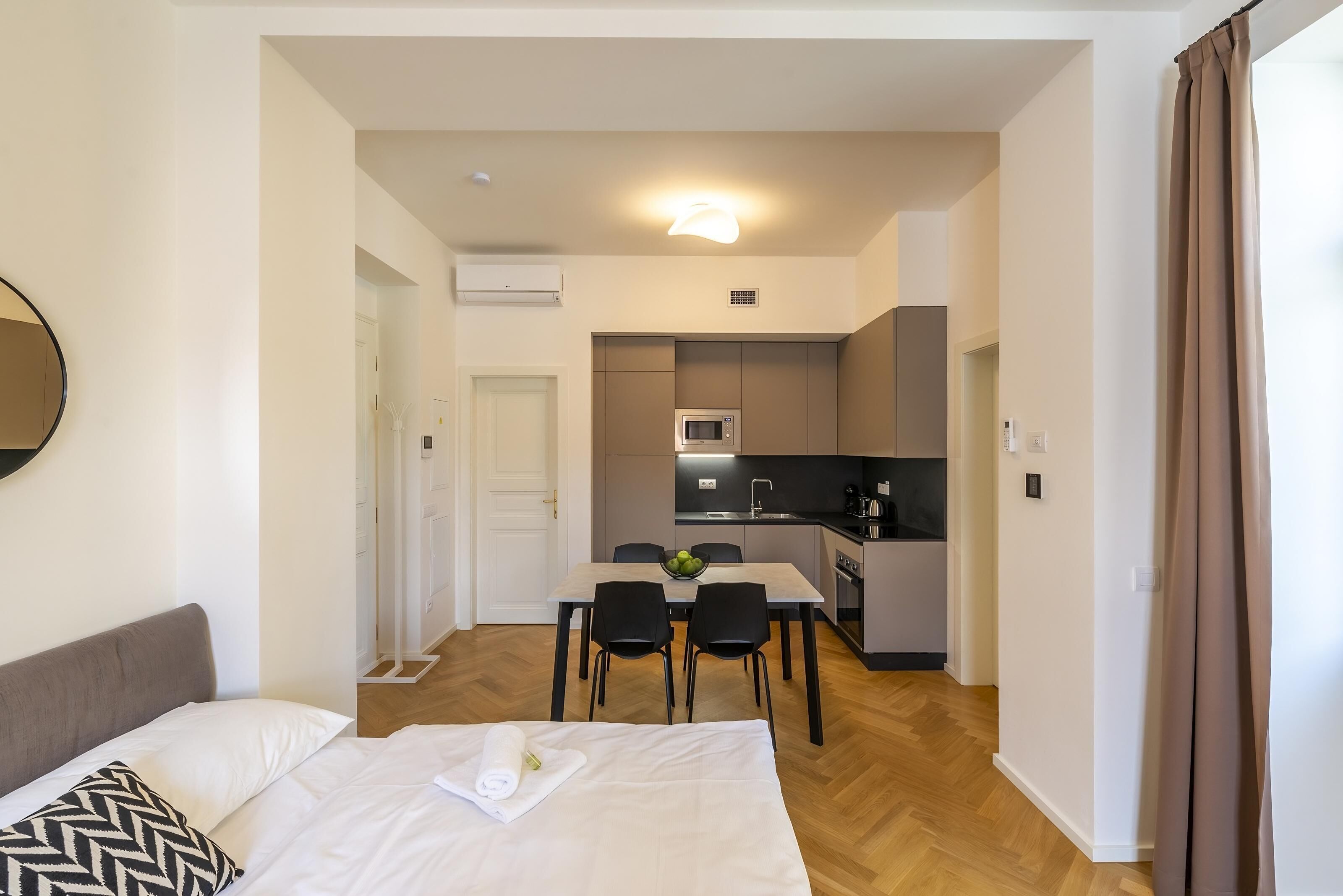 Una habitación de hotel moderna con una cama, una mesa de comedor con sillas, una zona de cocina con microondas y un baño visible a través de una puerta abierta.