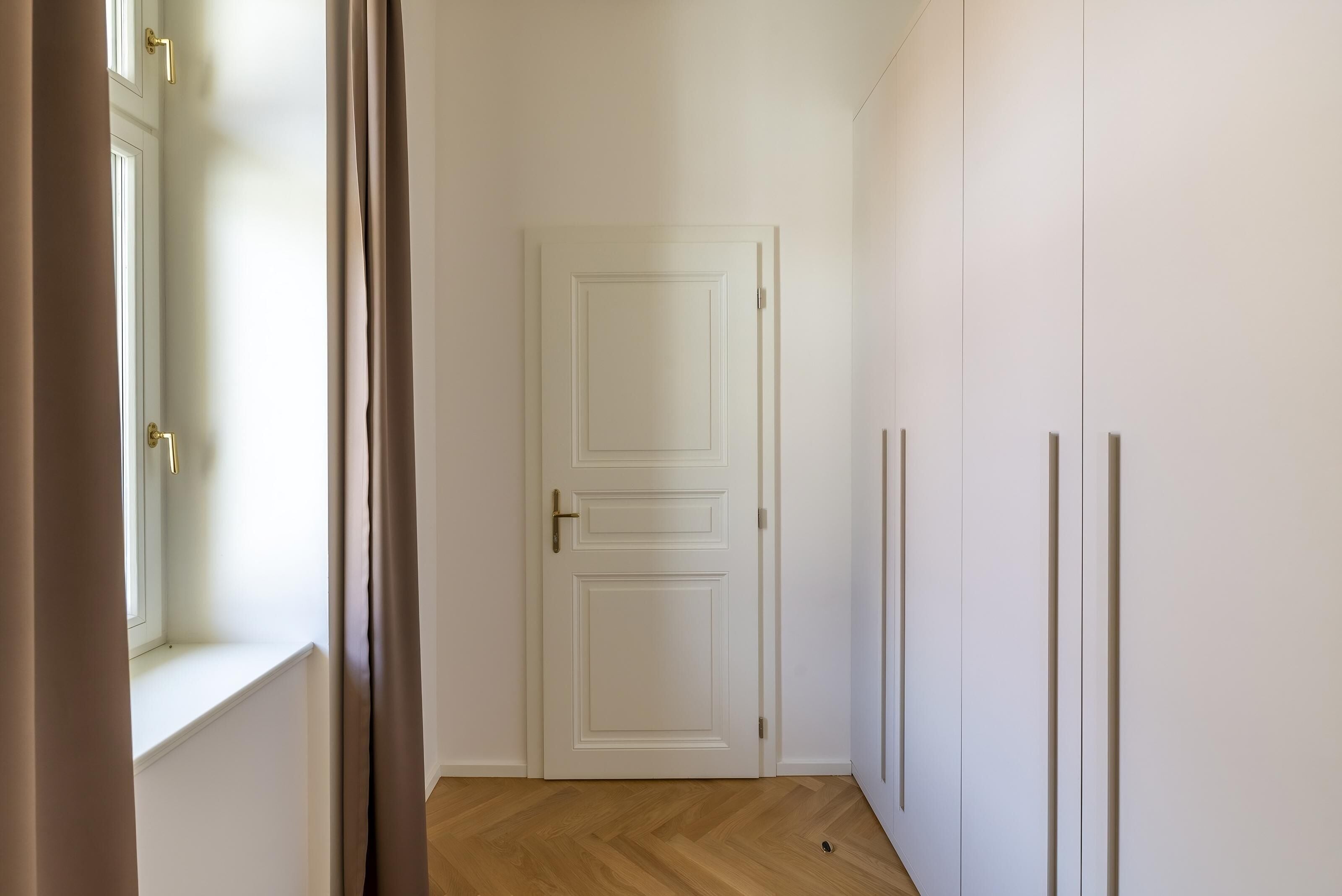 Un pasillo estrecho con una puerta blanca, una ventana con cortinas y suelo de madera.