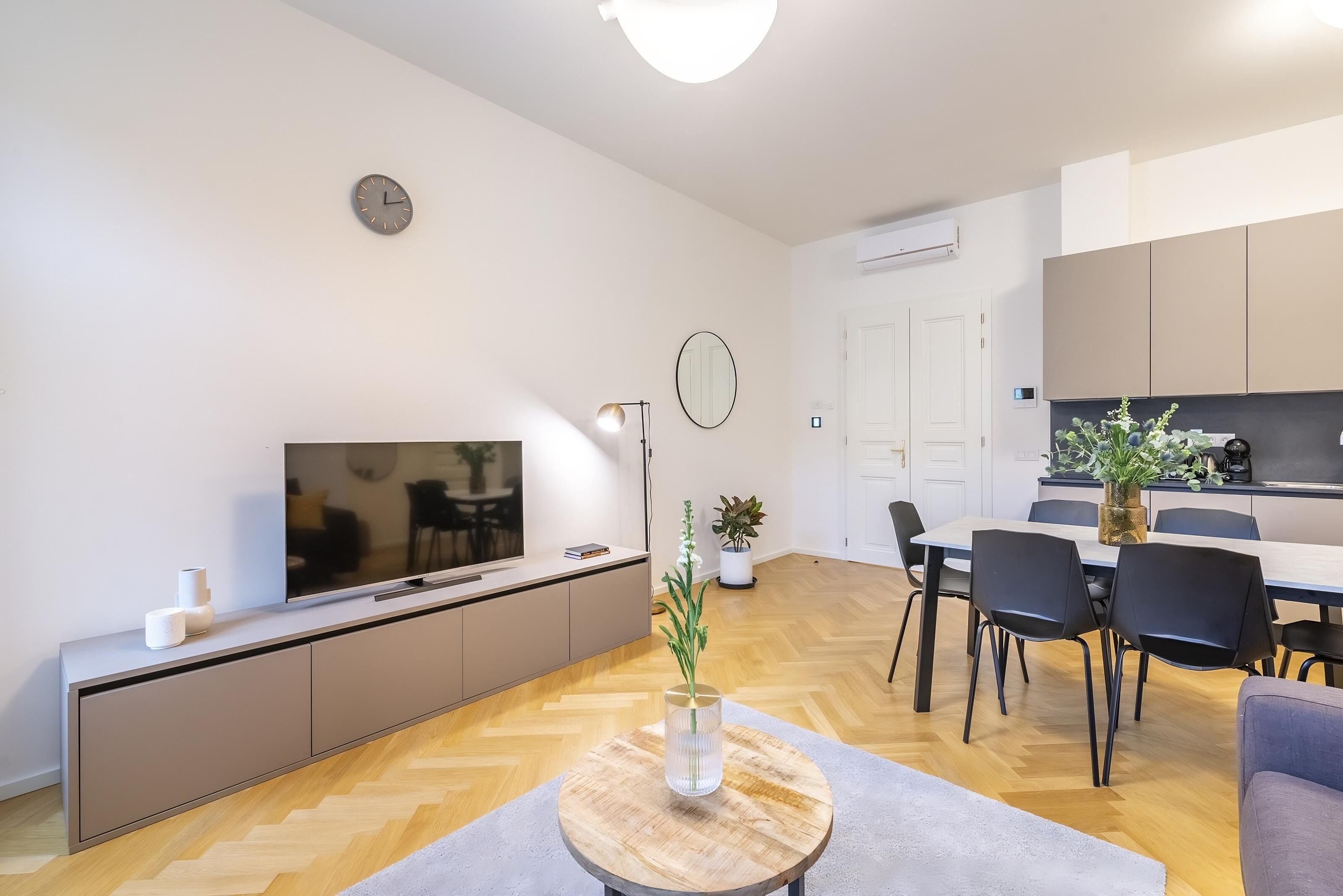 Uma sala de estar moderna com uma TV de tela plana, uma mesa de centro redonda de madeira e uma área de jantar, com uma cozinha ao fundo.