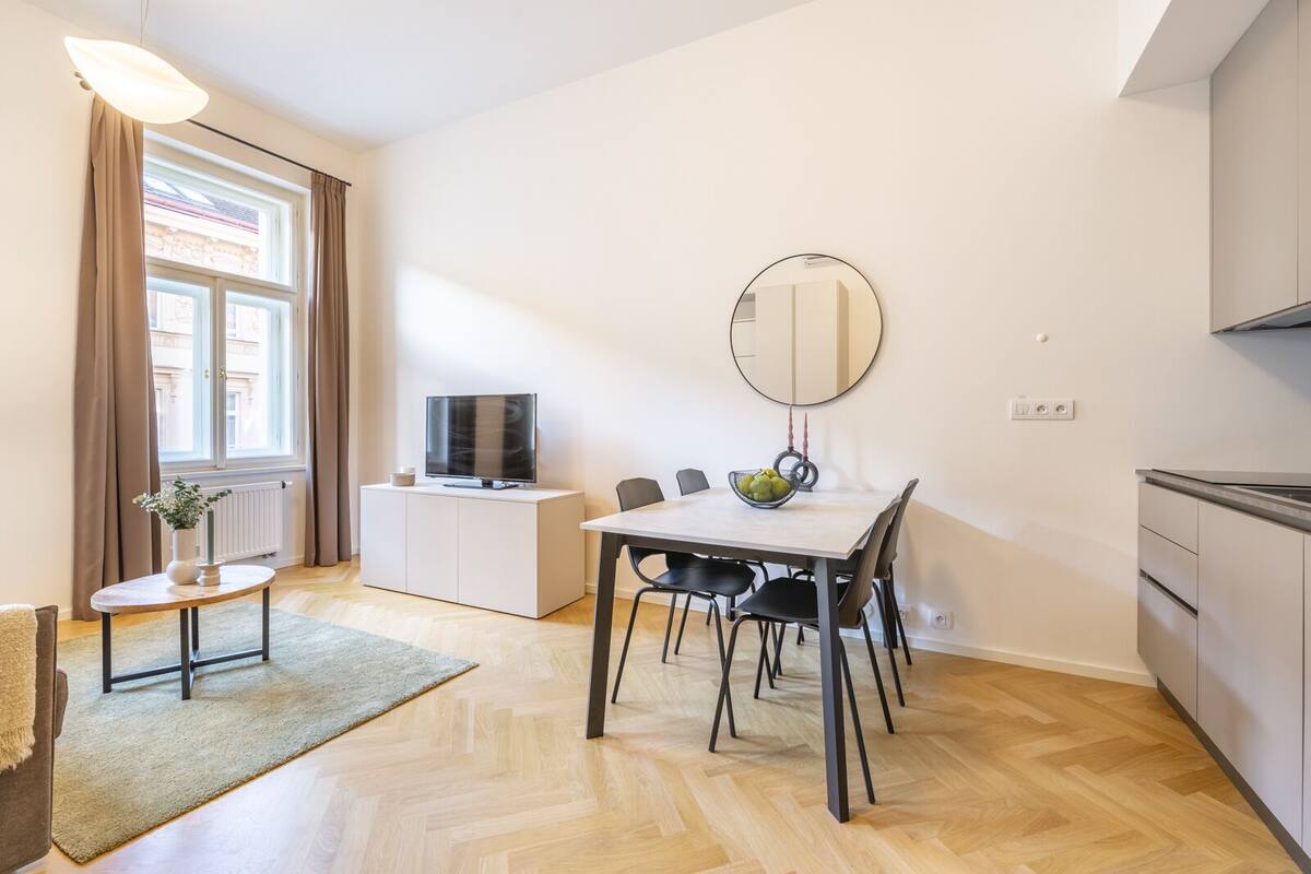 A modern living room with a dining table, chairs, a TV on a cabinet, a large mirror, and a window with curtains.