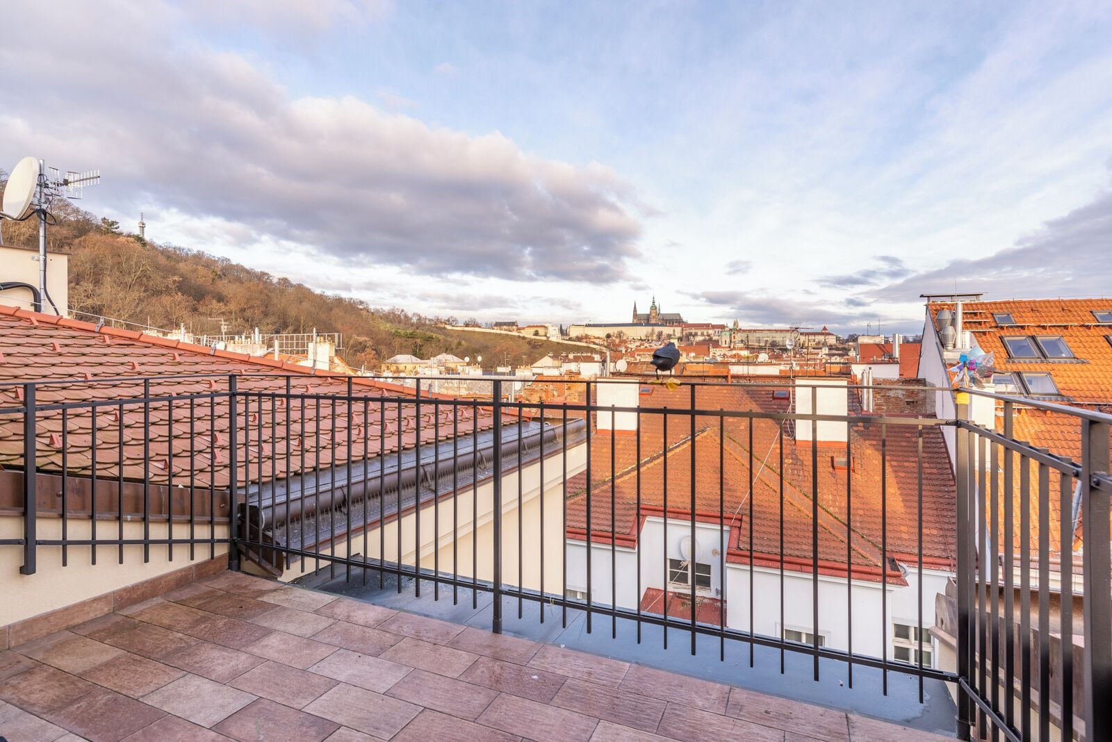 Una terraza en la azotea con barandilla de metal, vista de un paisaje urbano y una torre distante.