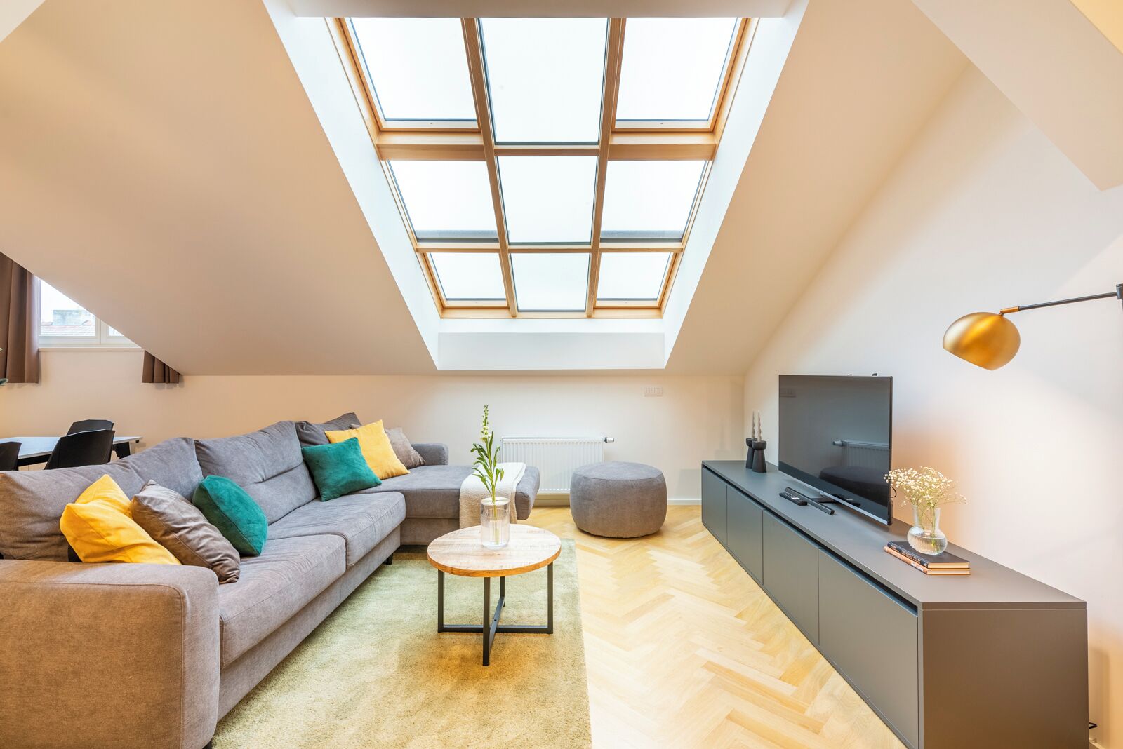 Moderni loft-tyylinen huoneisto, jossa on kattovalokuvio, harmaa sohva, pyöreä sohvapöytä, harmaa jalkarahi, televisio, joka on sijoitettu kaappiin, ja lattiavalaisin.