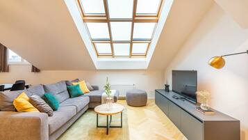 Moderni loft-tyylinen huoneisto, jossa on kattovalokuvio, harmaa sohva, pyöreä sohvapöytä, harmaa jalkarahi, televisio, joka on sijoitettu kaappiin, ja lattiavalaisin.