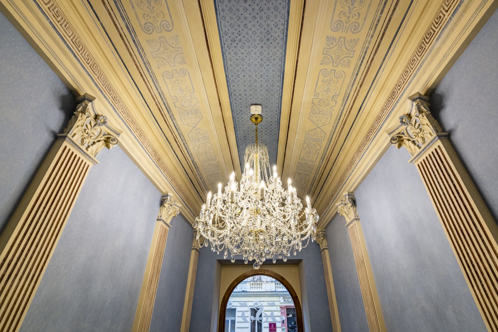 An ornate interior with a chandelier, classical columns, and detailed ceiling patterns.