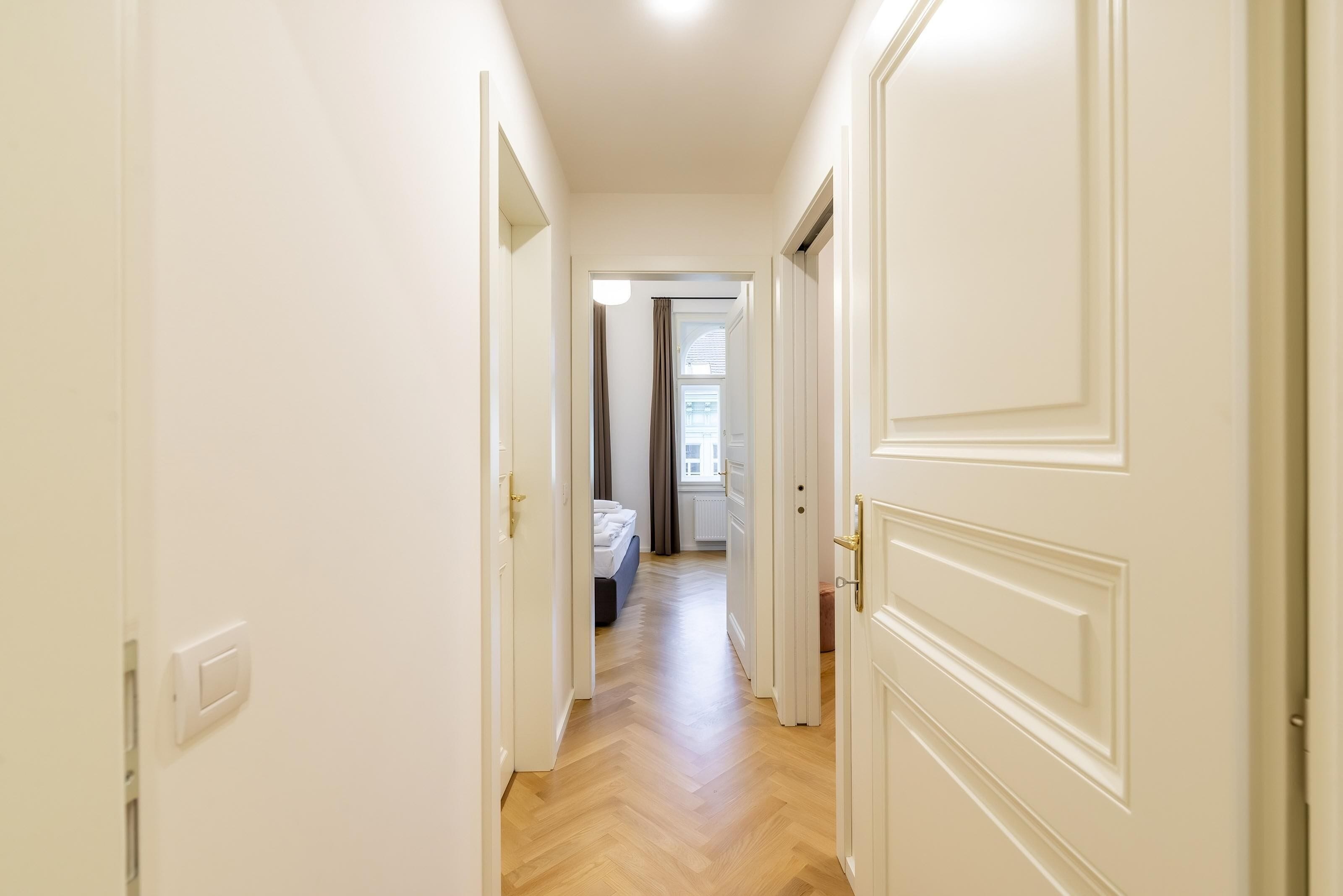 Ein enger Flur mit weißen Türen, die in ein Schlafzimmer mit Bett und Fenster führen.