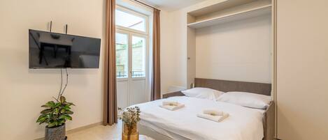 A modern hotel room with a bed, a TV mounted on the wall, a potted plant, and a small table.