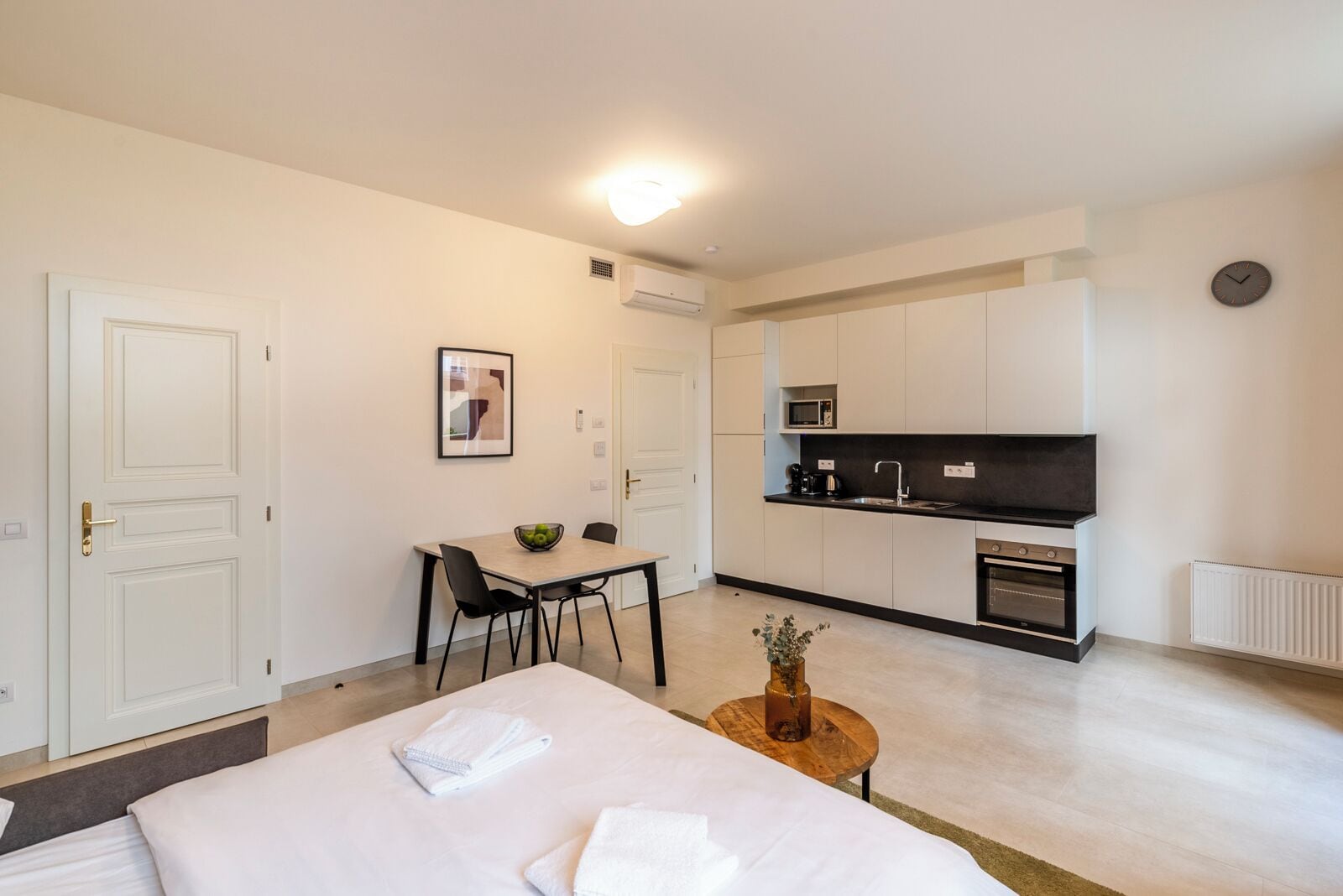Une chambre d’hôtel moderne comprenant un lit, une petite table à manger avec des chaises, un coin cuisine équipé d’un four à micro-ondes et d’une cuisinière, ainsi qu’une plante en pot.