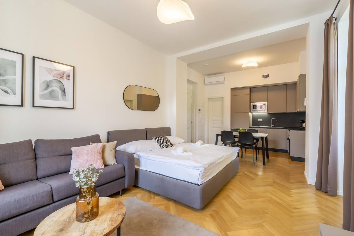 A modern living room with a grey sofa, a round coffee table, and a bed with white linens. There is a kitchen area with a microwave and a dining table with chairs.