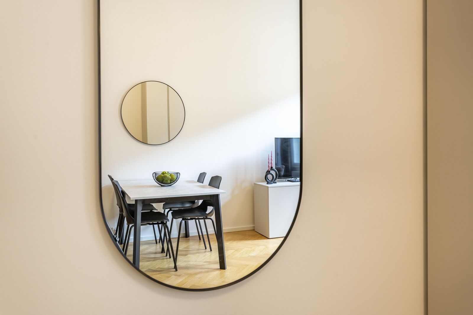 A dining area with a round mirror on the wall, a table with chairs, and a bowl of fruit.