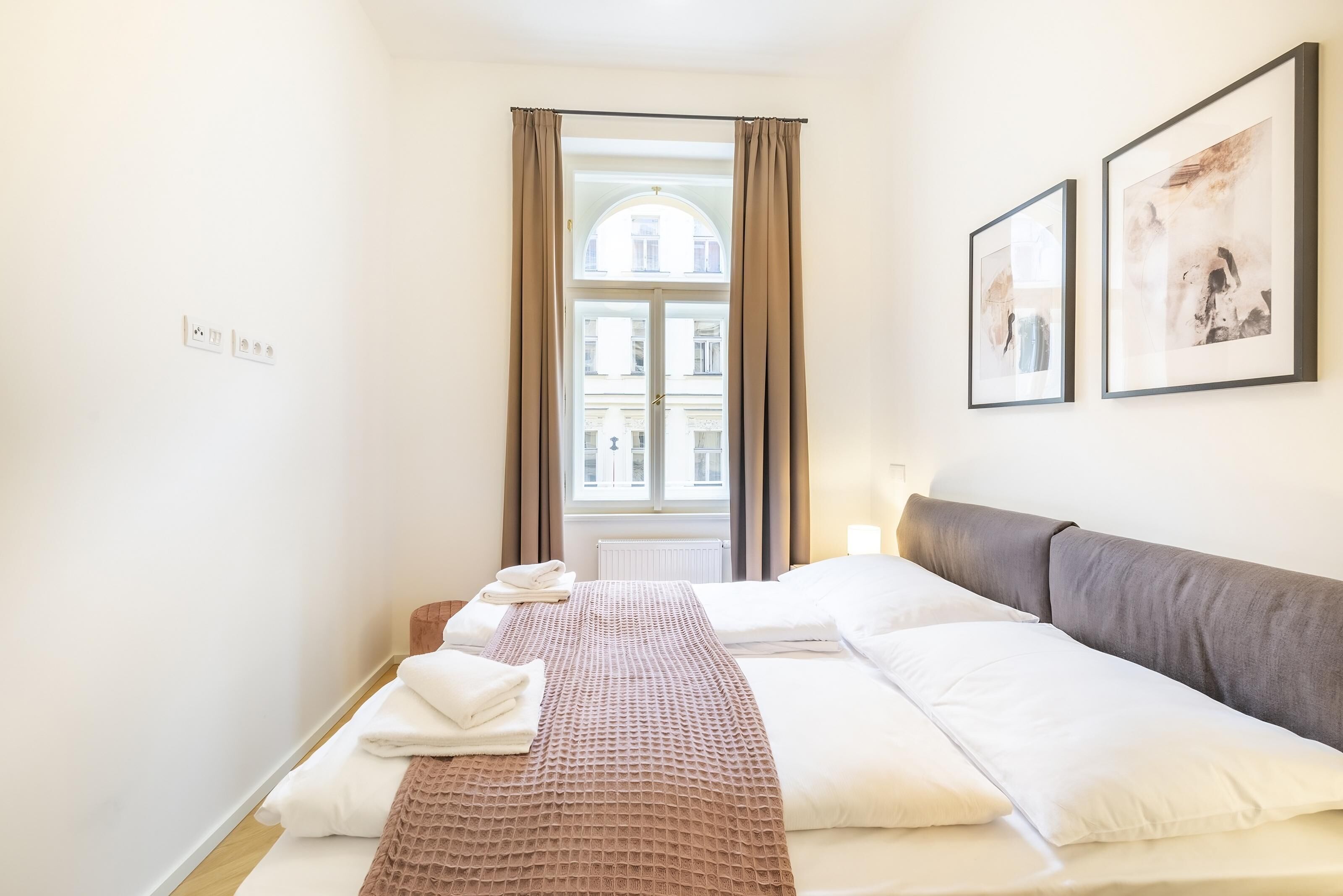 Une chambre d’hôtel avec un lit, une fenêtre avec des rideaux, une lampe fixée au mur et deux tableaux encadrés.