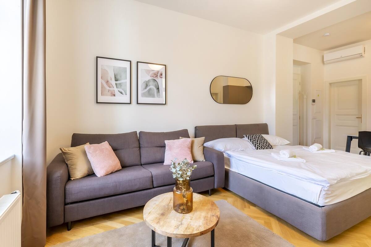 A modern bedroom with a grey sofa, a bed with white linens, a round wooden table with a vase, and two framed artworks on the wall.