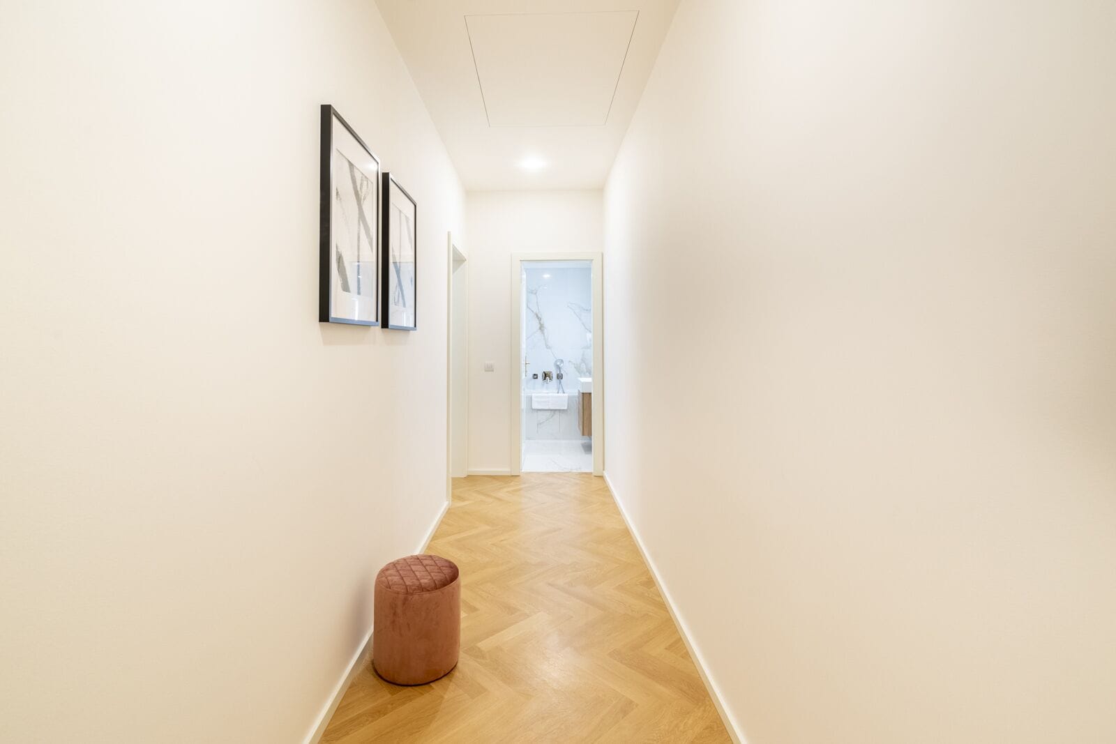 A narrow hallway with a wooden floor, a pink ottoman, and framed artwork on the walls.
