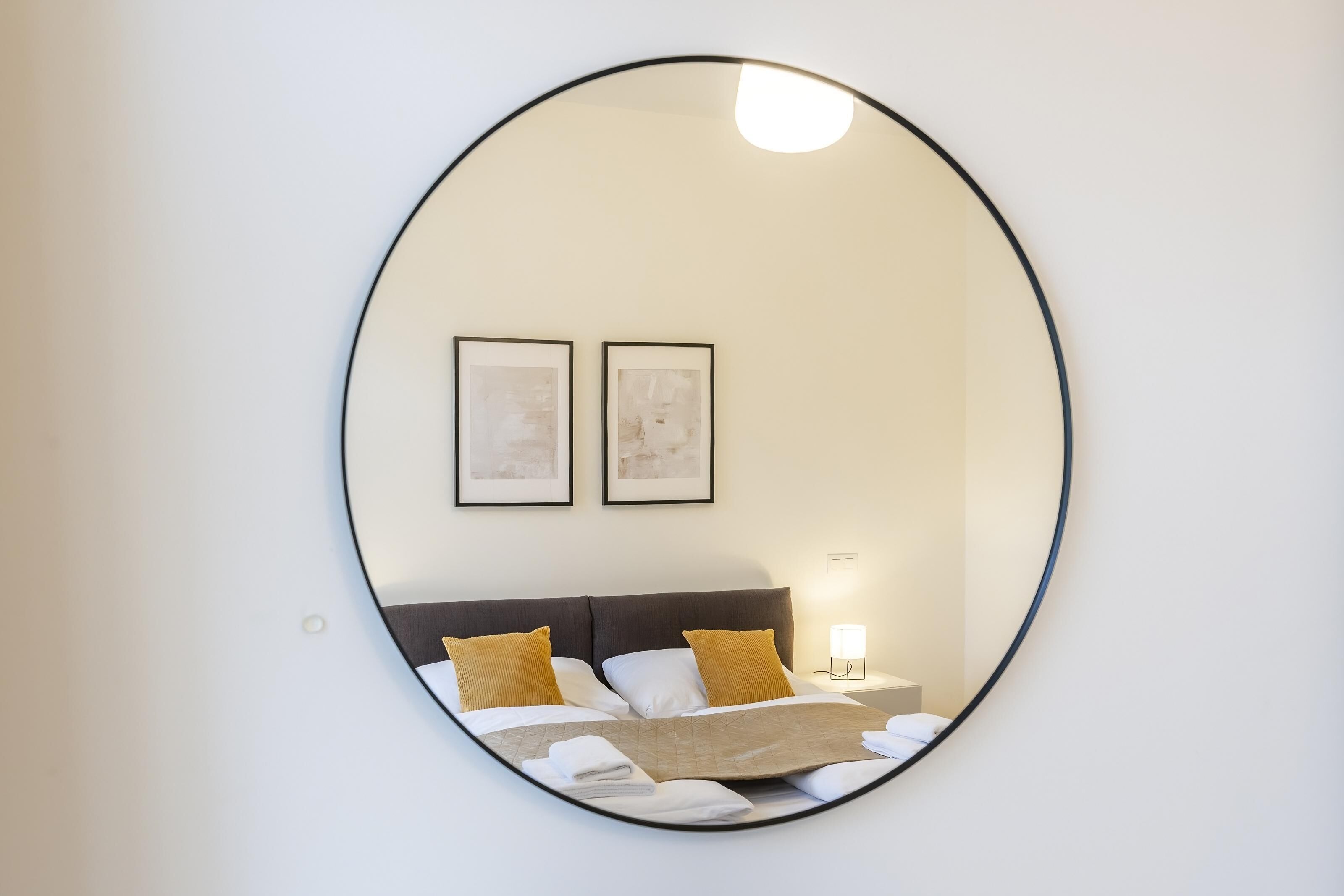 A circular mirror reflecting a neatly made bed with two pillows, a bedside table with a lamp, and two framed pictures on the wall.
