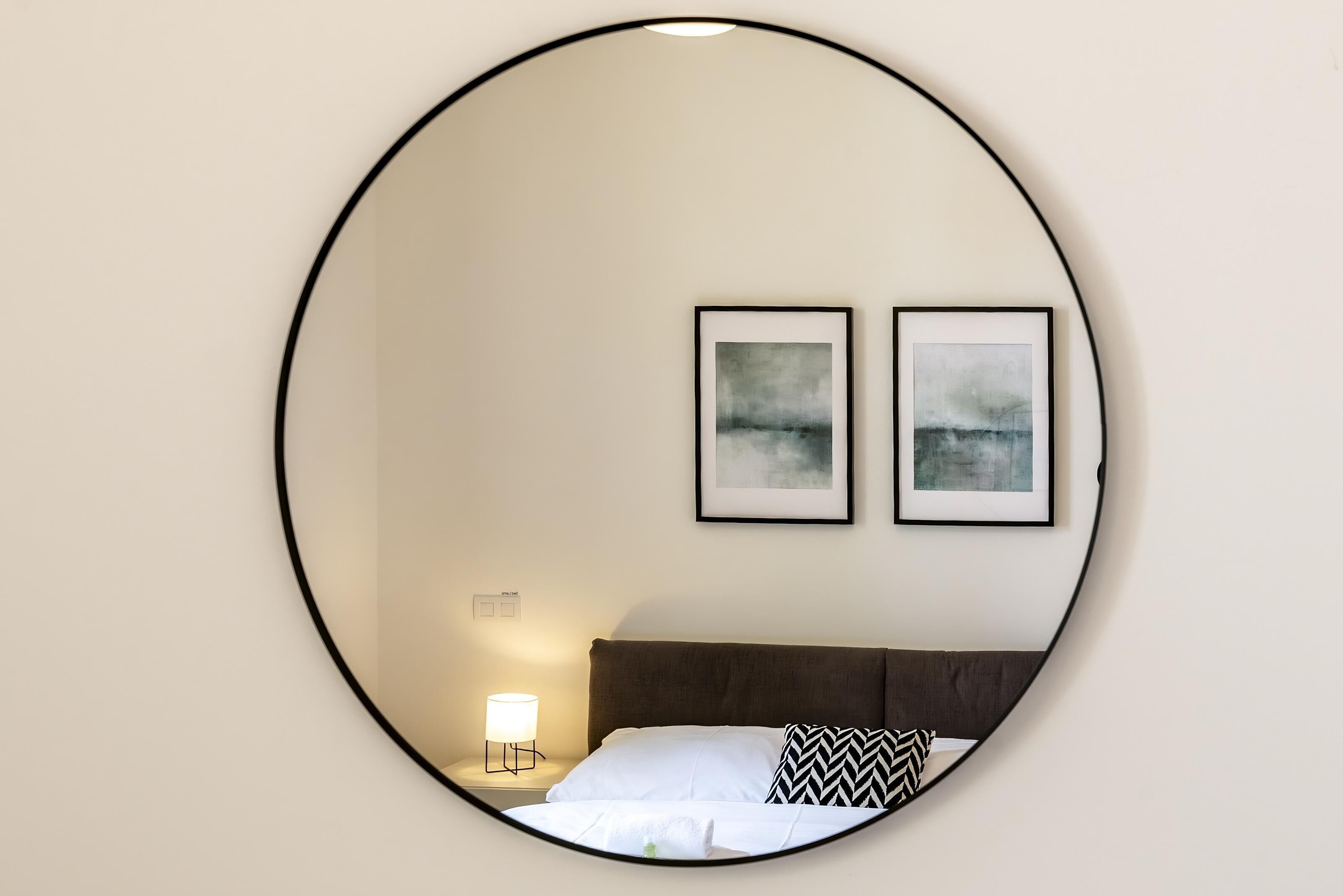 Un espejo circular refleja una cama con ropa de cama blanca y un almoháque con diseño blanco y negro, una lámpara de noche, y dos cuadros abstractos enmarcados en la pared.