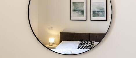 Un espejo circular refleja una cama con ropa de cama blanca y un almoháque con diseño blanco y negro, una lámpara de noche, y dos cuadros abstractos enmarcados en la pared.