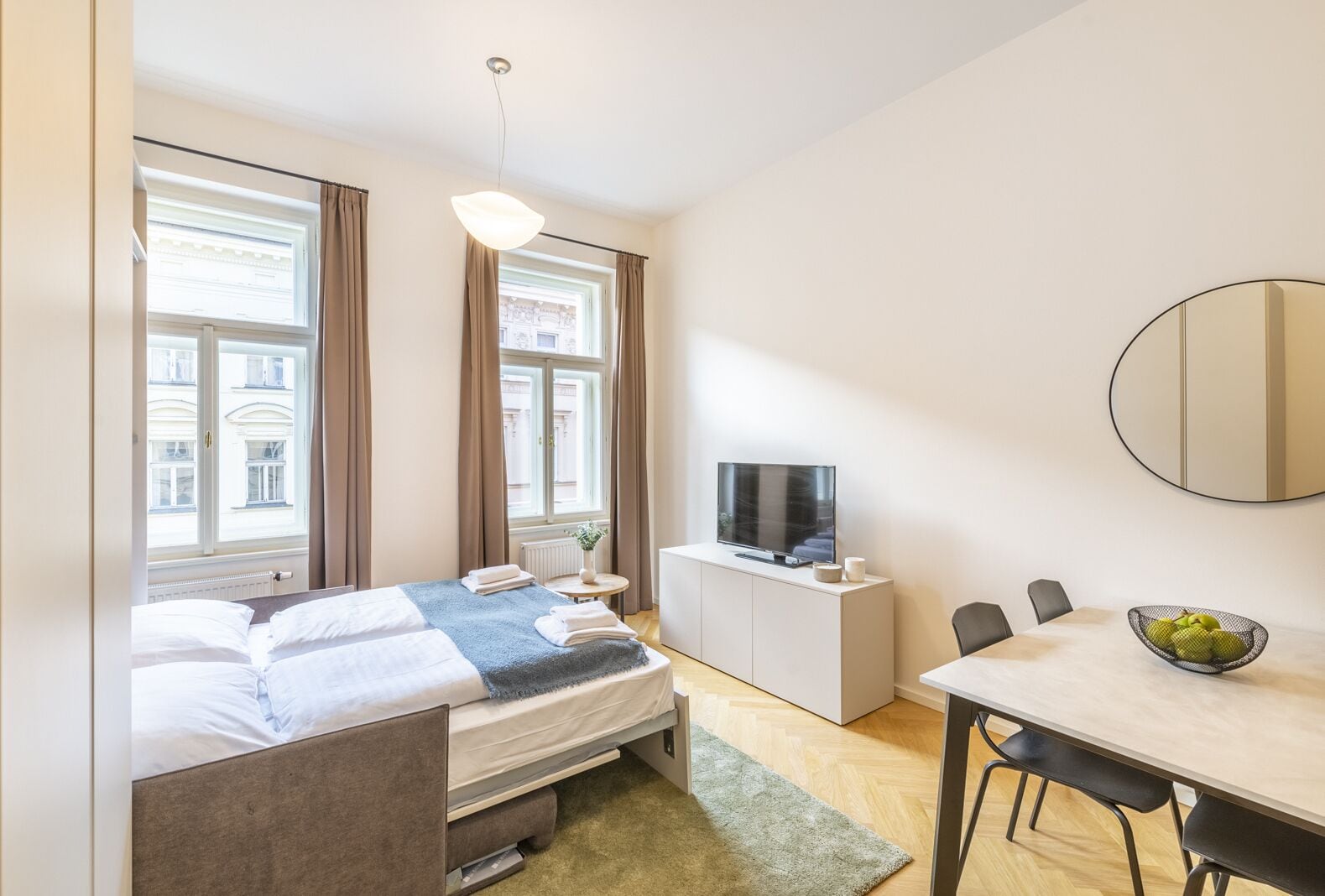 Een hotelkamer met een bed, een televisie, een eettafel met stoelen en een groot raam met gordijnen.