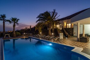 A swimming pool with lounge chairs, palm trees, and a modern building with a stone staircase. - Villa Rodea Cinco by Villa Plus (Playa Blanca)