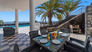 Un área de comedor al aire libre techada con piso a cuadros, muebles de mimbre y una mesa para dos personas con platos, vasos y un frutero.