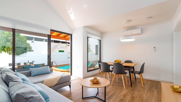 A modern living room with a grey sofa, a small round table, and a dining area with a wooden table and black chairs. There is a large window providing a view of a swimming pool and greenery outside. - Villa Famara by Villa Plus (Tías)