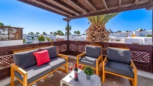 Una terraza con mobiliario de madera, una palmera y vistas a edificios blancos y al mar.