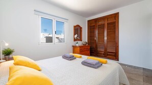 Un dormitorio con una cama, dos almohadas amarillas, una mesita de noche con lámpara, un aparador, un espejo y una ventana con vistas a edificios.