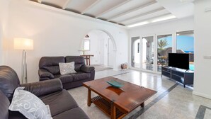 A spacious living room with a leather sofa, a wooden coffee table, and a floor lamp. There is a large window with a view of the outdoors. - Villa Nino by Villa Plus (Puerto del Carmen)