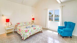 A bedroom with a bed, a blue armchair, a window with a view, and two bedside lamps. - Villa Nino by Villa Plus (Puerto del Carmen)