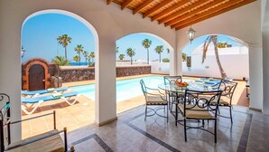 A covered outdoor area with a pool, seating, and a view of palm trees and the sea. - Villa Nino by Villa Plus (Puerto del Carmen)