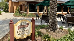 Un bar-restaurant avec une terrasse, une enseigne indiquant qu’il s’agit d’un magasin d’articles de golf, et un bâtiment doté d’un toit en tuiles et bordé de palmiers.