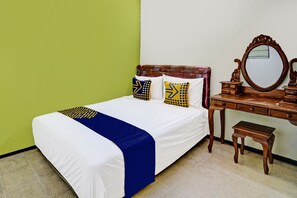 Un lit bien fait, recouvert d’une courtepointe blanche et bleue, deux oreillers aux motifs géométriques, une commode en bois avec un miroir et un petit tabouret en bois.