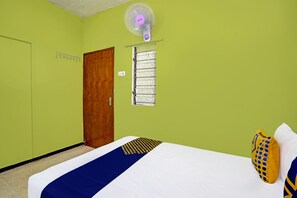 Une pièce avec un lit, une porte en bois, une fenêtre avec des stores et un ventilateur fixé au mur.
