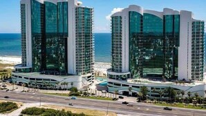 Dos edificios altos con fachadas de vidrio reflectante, una estructura curva y vista a la playa.