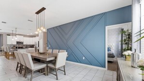 A modern kitchen with white cabinets, a dining area with a wooden table and chairs, and a blue accent wall with a geometric pattern. - Champions Gate 4 BD, Themed, Private Pool 1542 (Davenport)