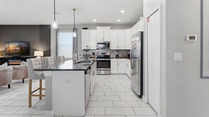 A modern kitchen with white cabinetry, a central island, stainless steel appliances, and a dining area with a gray upholstered table and chairs. - Champions Gate 4 BD, Themed, Private Pool 1542 (Davenport)