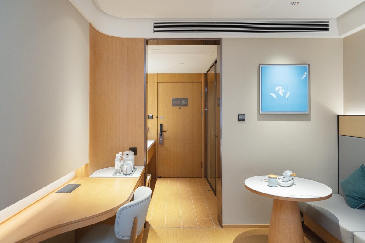 Habitación de hotel con puerta de madera, una mesita con tetera y tazas, un sofá con cojín y una obra de arte colgada en la pared.