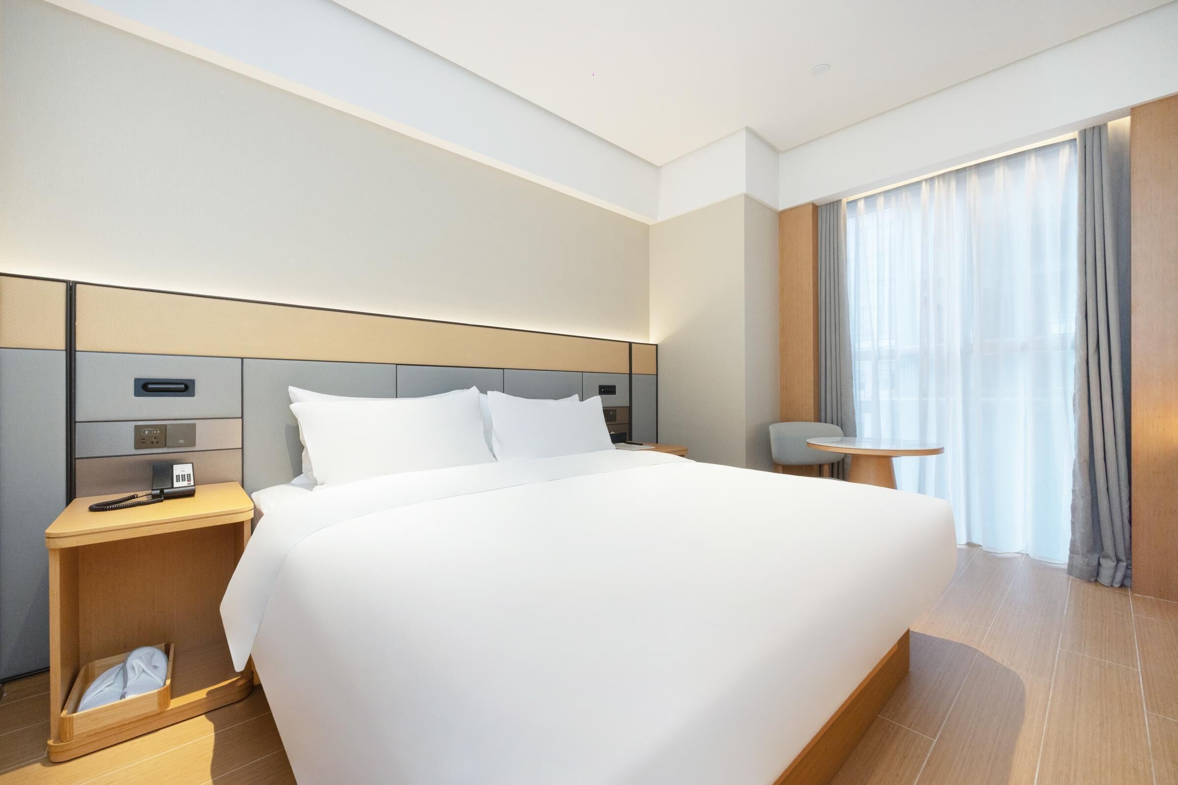 Una habitación de hotel moderna con una cama grande, una mesita de noche con lámpara, una pequeña mesa y una ventana con cortinas.