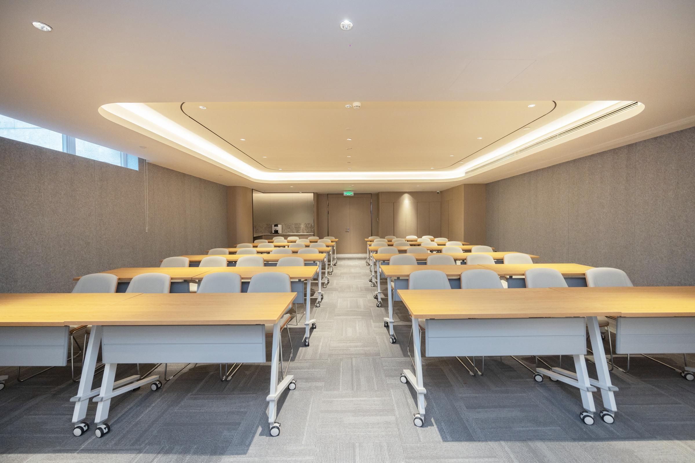 A conference room with rows of tables and chairs, a carpeted floor, and a ceiling with recessed lighting.