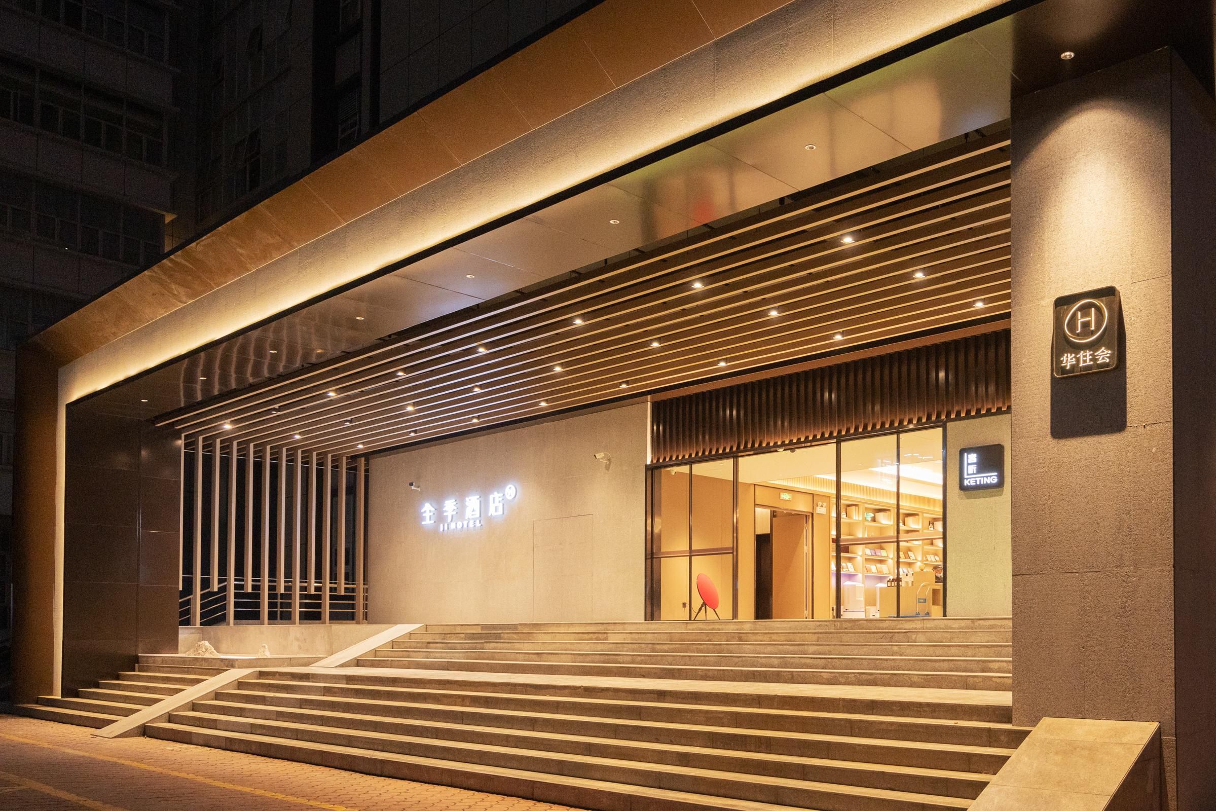 Entrada de hotel com design moderno, com uma escadaria que leva à porta, uma placa com o nome do hotel e um logotipo na parede.