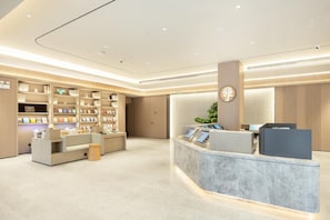 Un hall d’hôtel moderne avec un comptoir d’accueil, des étagères remplies de livres et d’objets décoratifs, et une grande horloge au mur.
