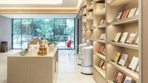 Eine moderne Bibliothek mit großen Fenstern, einem zentralen Schreibtisch mit Büchern und Kisten sowie Bücherregalen.