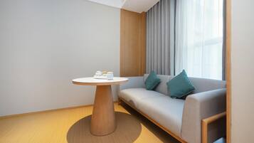 Una habitación de hotel moderna con una mesa redonda de madera, un sofá gris con cojines color esmeralda y un ventanal con cortinas de encaje.