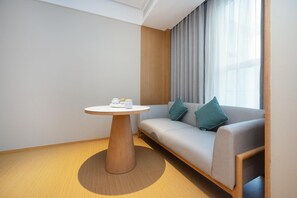 Una habitación de hotel moderna con una mesa redonda de madera, un sofá gris con cojines color esmeralda y un ventanal con cortinas de encaje.