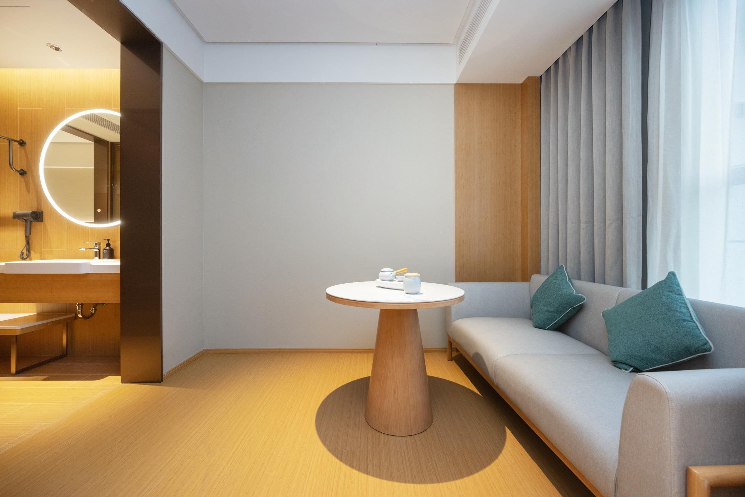A modern hotel room with a round wooden table, a sofa with teal cushions, a round mirror with lights, and a large window with curtains.