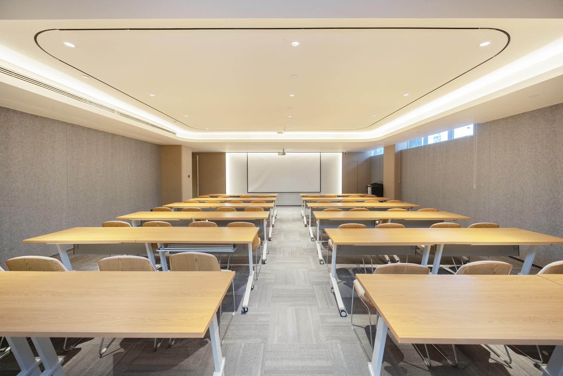 Uma sala de conferências com fileiras de mesas e cadeiras, piso coberto por carpete e uma tela de projeção na frente.