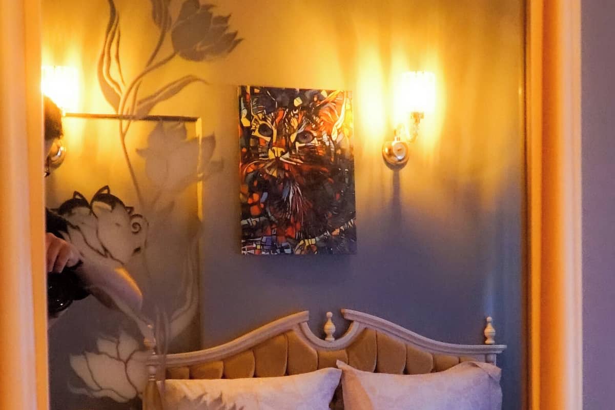 A bedroom with a bed, two wall sconces, a painting, and a mirror reflecting the room.