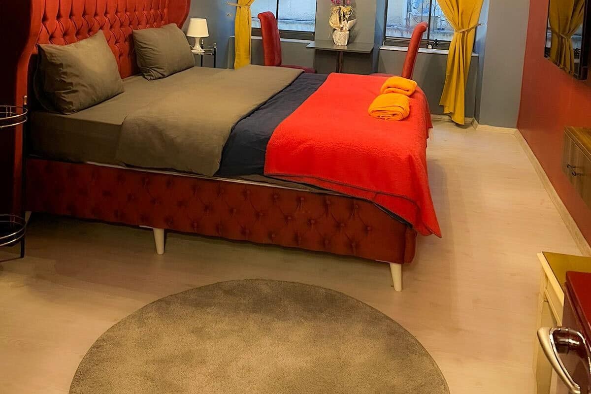 A bedroom with a red upholstered bed, a round rug, and yellow curtains.