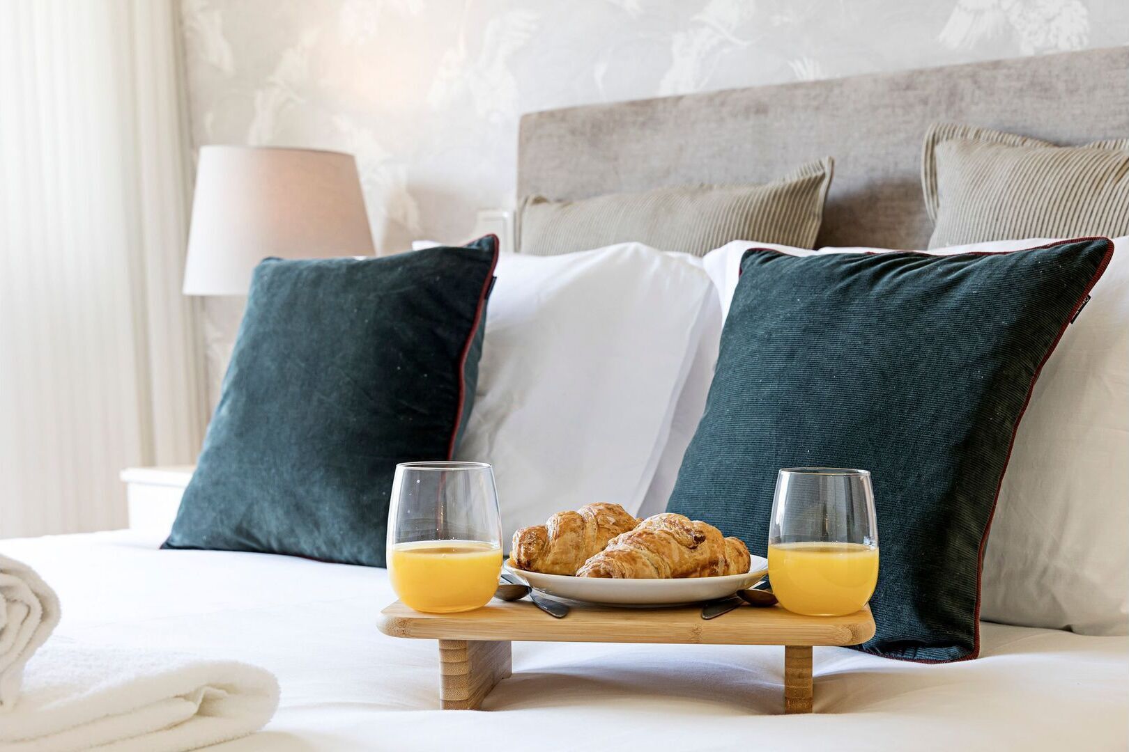 Een net opgemaakt bed met kussens, een bord croissants en twee glazen sinaasappelsap op een houten dienblad.
