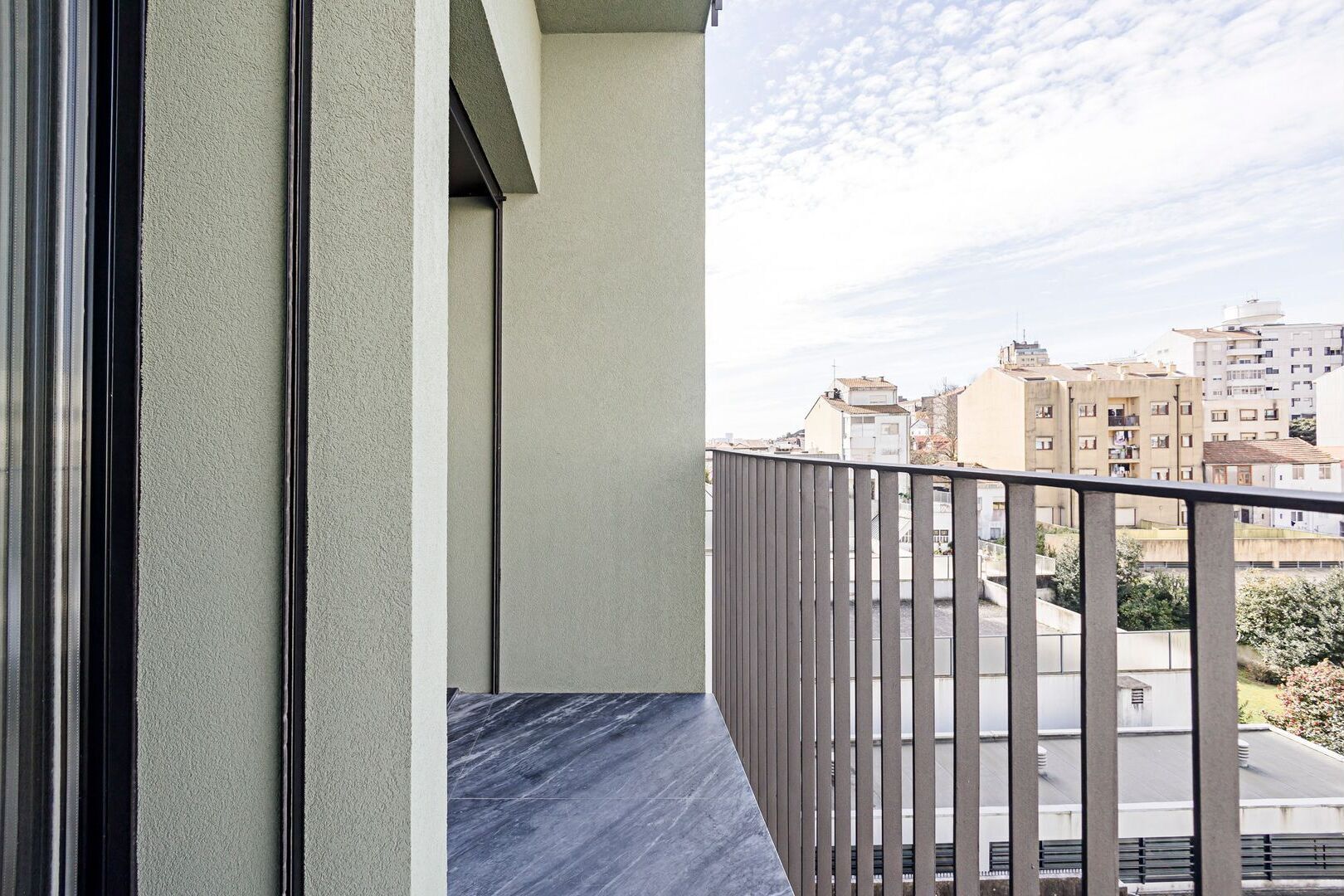 Een balkon met een metalen hekwerk, uitzicht op woongebouwen en een heldere lucht.