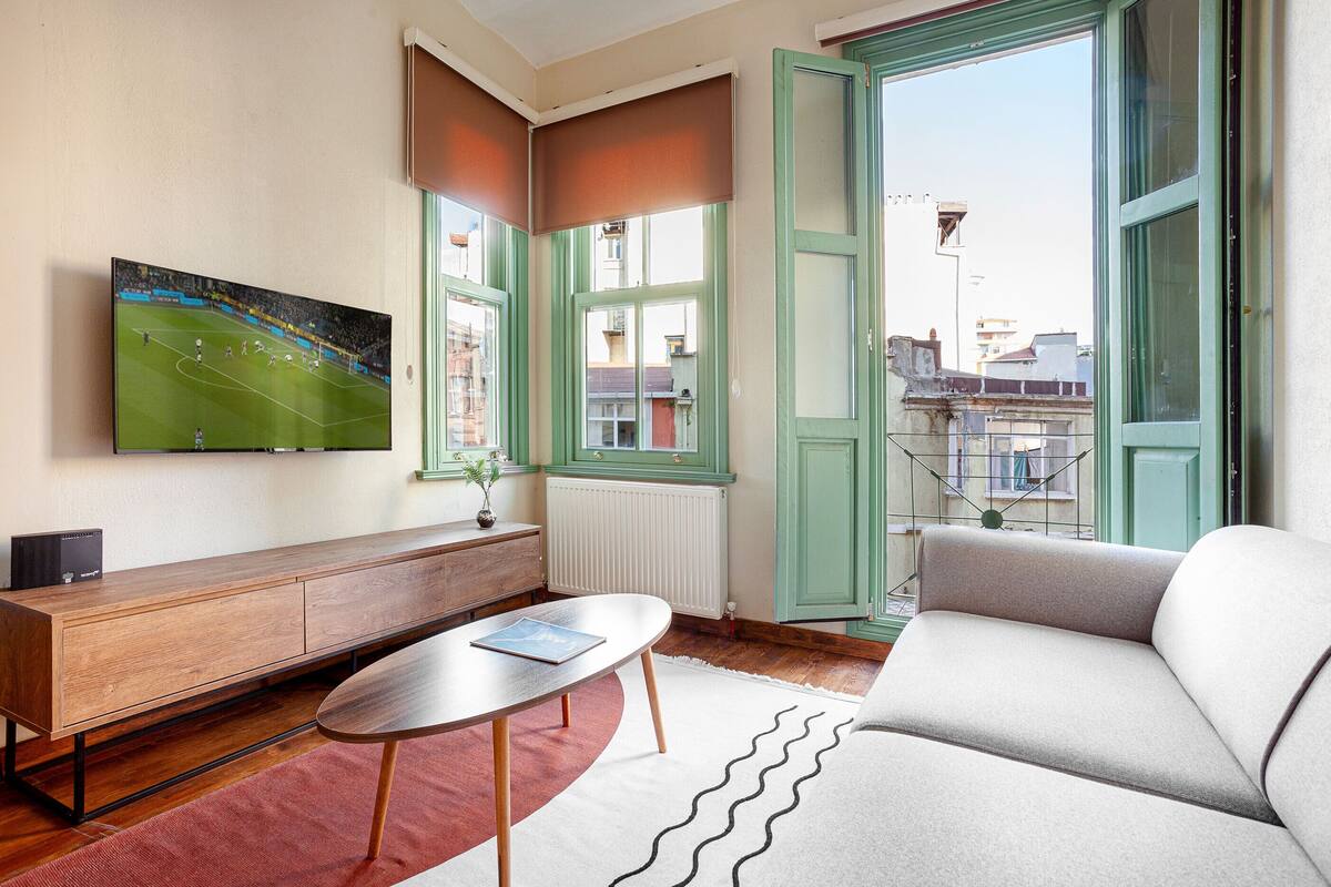 A living room with a sofa, a coffee table, a TV mounted on the wall, a wooden cabinet, and a view of the outside through a window.